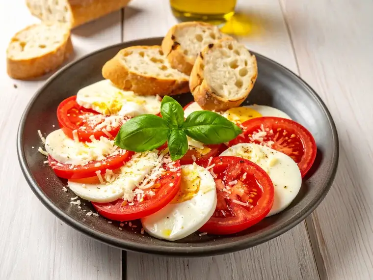 Caprese-Salat mit Tomaten, Reibekäse und Ei auf einem Teller, dazu Baguette